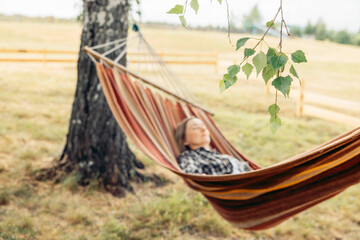 A young Caucasian woman with long brown hair relaxes in a colorful hammock under a tree in a rural setting. The scene conveys a sense of tranquility and escape.