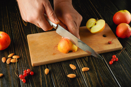 Hands are slicing a red apple on a wooden cutting board. The scene includes scattered almonds and red currants, creating a healthy snack setup in a cozy kitchen