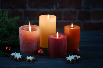 Four orange and red candles bringing warm light in the dark night for Christmas, Advent or New Year, some cookies, baubles and fir branches against a brick wall, selected focus