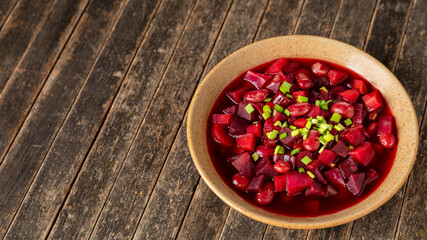 Homemade, vegan beet borscht (in Ukrainian style) topped with scallions on rustic wooden table with copy space. Soup made of beetroot, beans, cabbage, carrot, and celery stalks.  