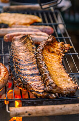 Vertical close-up of two golden pork ribs slowly roasting on a barbecue rack with sausages and chicken fillets in the background.