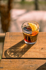 A refreshing glass of vermouth with an orange slice and ice on a wooden picnic table outdoors on a sunny day, casting a long shadow on the table.