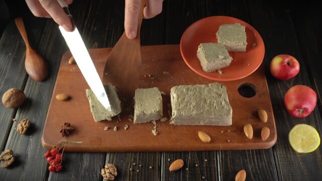 A person slices a block of nut-based dessert or halva on a wooden cutting board surrounded by fresh fruits and nuts. The warm kitchen invites a sense of homemade joy and creativity.