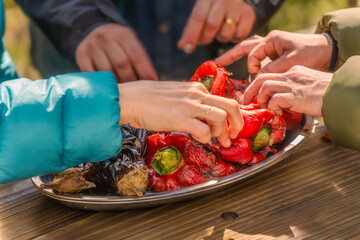 Group of women's hands peeling roasted red peppers and eggplants on a tray on a wooden table after being roasted on an outdoor barbecue.
