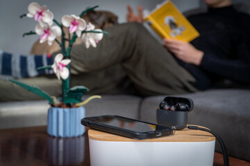 Smartphone and headphones set aside in the foreground while a man reads a book in the background. Concept of digital detox, disconnecting from technology, and returning to analog leisure