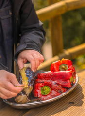 Women's hands peeling roasted eggplants next to red peppers on a tray on a wooden table, after being grilled on an outdoor barbecue.