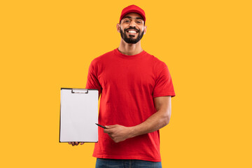 A man with a beard wears a red shirt and cap while he stands against a vivid yellow background. He...