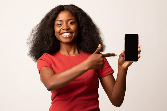 Cheerful African American woman points at a blank smartphone screen, promoting a new application or website. She stands against a plain white background, conveying a friendly message about technology.