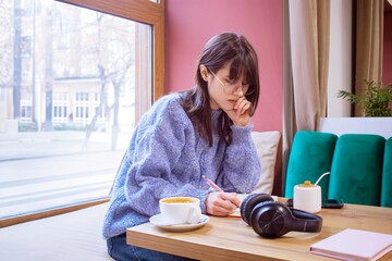 Young woman writing in a notebook studying at a cafe table. Concentrated young woman wearing glasses in a cozy cafe setting.