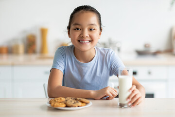 A joyful asian girl sits in a kitchen, smiling at the camera while enjoying cookies and milk. The table has a plate of freshly baked biscuits, showcasing a moment of happiness.