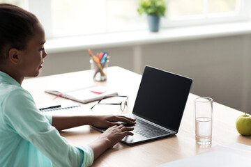 A teenage African American girl types on her laptop with an empty screen while studying at home. She is focused on her work in a bright room, surrounded by school supplies and a refreshing drink.
