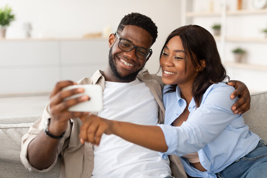 Young black couple sitting on their couch, smiling and using a cellphone together. The man embraces the woman as she points at the screen, both joyous.