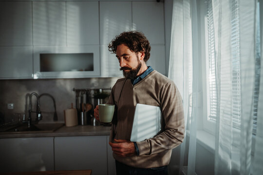 Man thinking holding coffee and laptop in kitchen - Powered by Adobe