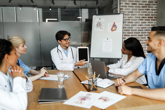 Chief physician holding digital tablet with blank screen while having brainstorming or medical training with international group of doctors, mockup, modern technologies and healthcare cocnept