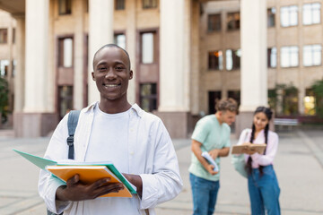 A joyful student holding multiple books poses in front of a university. Two classmates are visible in the background discussing their study materials together.