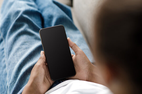 Young black girl sits comfortably on a sofa at home, focused on her smartphone. She types, watches videos, or searches for information, staying connected during covid-19 quarantine.