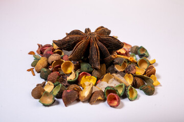 A macro photograph of a single dried Star Anise fruit resting on a pile of smaller, brightly colorized botanical seed capsules and clear crystalline material on a bright white backgr