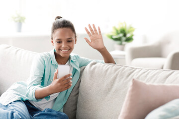 A teenage girl happily waves at her webcam during a video call in her cozy living room. She is staying home, following social distancing guidelines due to the Covid-19 pandemic.