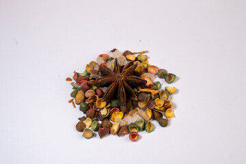 A macro photograph of a single dried Star Anise fruit resting on a pile of smaller, brightly colorized botanical seed capsules and clear crystalline material on a bright white backgr