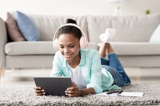 Young girl with headphones is happily studying online while lying on the floor in her living room. She engages with her tablet and has notes nearby, showcasing her dedication to learning at home. - Powered by Adobe