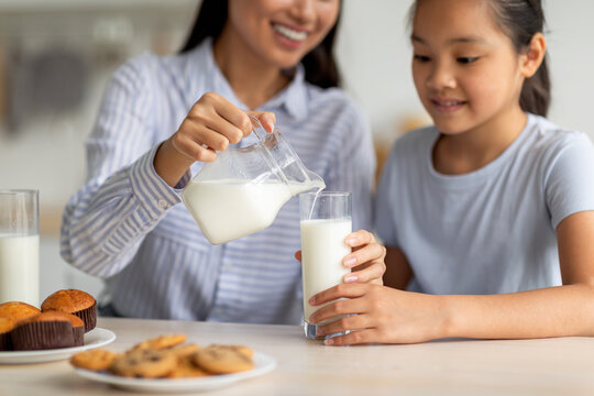 An Asian mother serves fresh milk to her teen daughter in a cozy kitchen. They enjoy breakfast together, highlighting a moment of health and family bonding over food and drinks.