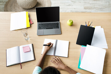An afro American girl student is typing notes in a notebook while working at her desk at home. She is focused on her studies with a laptop and various materials nearby.