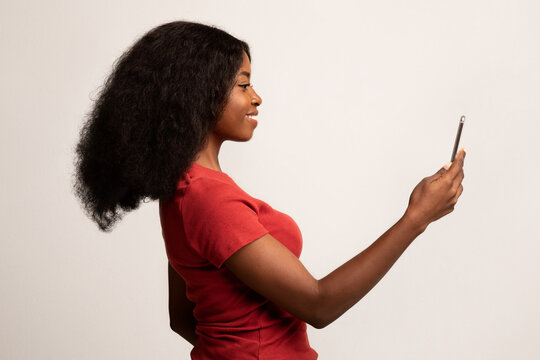 A young Black woman stands alone against a white background, smiling as she uses a face recognition app on her smartphone.
