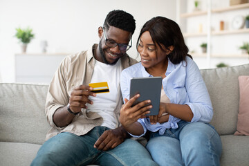 Millennial black couple sits comfortably on their sofa at home, exploring options on a tablet. The excited woman points at the screen while her partner holds a bank card, ready to purchase.