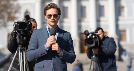 Male journalist in a stylish suit stands confidently with a microphone, surrounded by cameras, capturing a news event outdoors, showcasing the essence of modern journalism