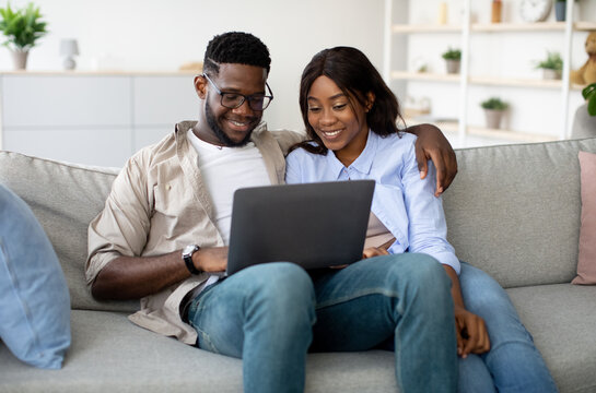 Joyful African American couple sits on a sofa at home, enjoying a movie together on a laptop. They cuddle closely, sharing smiles while browsing the internet during their weekend.