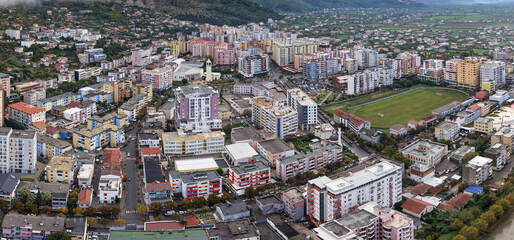 Obraz premium Aerial image of central Lezhë with colourful residential blocks, hillside homes, and the city stadium under cloudy skies.