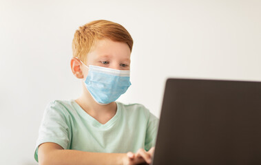 A redhead boy sits at a desk wearing a face mask while using a laptop for home-schooling. He focuses on typing, participating in online education during the coronavirus pandemic.
