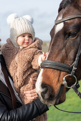 A little girl in a warm knitted hat and fluffy overalls meets a horse for the first time during hippotherapy at an equestrian club.