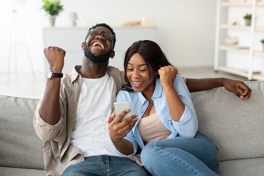 An emotional African American couple celebrates a recent online win while using their smartphone. They are on a comfortable couch, showing excitement with clenched fists and joyful expressions.