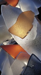 Bright sunlight illuminates layered orange and white glass fragments, creating a vivid abstract pattern of sharp shapes and strong contrast.
