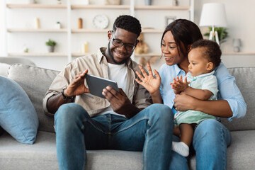 A smiling African American man, woman, and boy sit on a couch in their living room, waving at the tablet during a video call. They share moments with loved ones, creating joyful memories together.