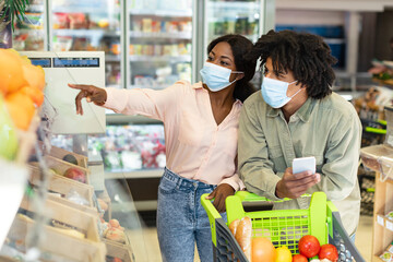 In a grocery store, a couple explores fresh produce while wearing face masks. The woman points out items of interest as the man holds a smartphone and cart.