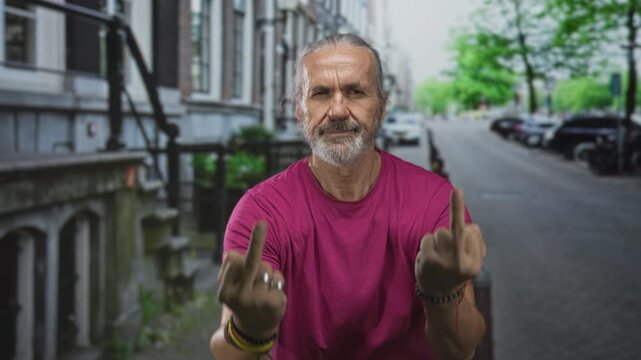 Man shows both middle fingers on a city street, wearing magenta shirt and long grey hair tied back with visible rings and bracelets; defiance.