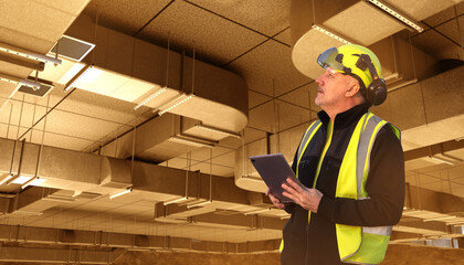 Building inspection specialist evaluates ceiling ventilation systems using a digital tablet, checking structural elements and airflow components in an industrial facility.
