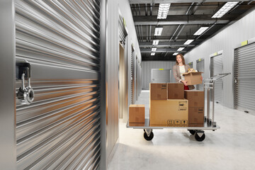 Storage facility customer transports packed boxes on a cart, organizing belongings and preparing items for placement in a secure rental unit corridor.