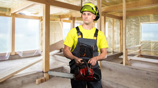 Construction worker holding a chainsaw inside a partially built wooden structure, wearing safety gear including a helmet, goggles, and overalls.