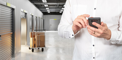 Storage facility manager uses a smartphone to control unit access and track stored goods while overseeing boxes and carts in a modern self-storage corridor.