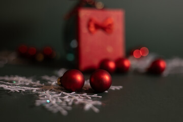 A festive red gift box with a bow surrounded by red Christmas ornaments and delicate white snowflake decorations on a dark green background.