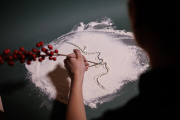 A creative moment where a person uses a berry branch to draw the outline of a girl’s face in a layer of flour, capturing an artistic, delicate scene filled with imagination and expressive detail.