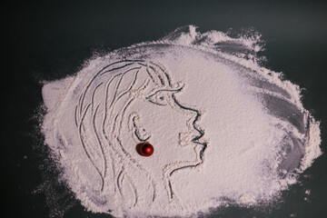 Artistic face illustration drawn in flour on a dark surface, accented with a small red Christmas ornament for a festive creative touch.