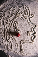 Artistic face illustration drawn in flour on a dark surface, accented with a small red Christmas ornament for a festive creative touch.