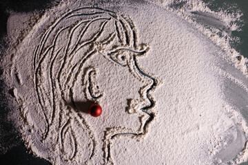 Artistic face illustration drawn in flour on a dark surface, accented with a small red Christmas ornament for a festive creative touch.