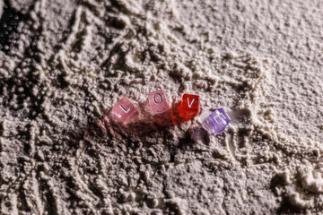 The word “LOVE” arranged from small cubes on a surface dusted with white flour, softly illuminated from the side by warm light, creating a cozy and delicate romantic atmosphere.