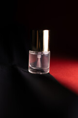 Elegant still-life of a clear nail polish bottle with a golden cap, placed on a dramatic black and red background with moody lighting that highlights its glossy, luxurious appearance.