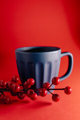 Festive still-life featuring a blue ceramic mug placed against a vibrant red background, accented with glossy red berries for a warm, seasonal holiday atmosphere.